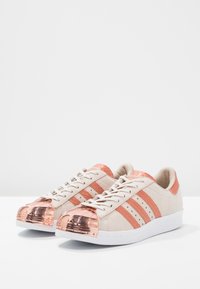Beige sneakers med roséguld accenter, med tre ränder, en blank tåhuvud och texturerad detaljer på ovandelen. Vit sula.