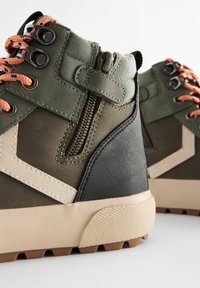 Des baskets montantes vertes présentent un design texturé avec des lacets orange, une fermeture éclair sur le côté et une semelle en caoutchouc robuste avec des motifs de traction.