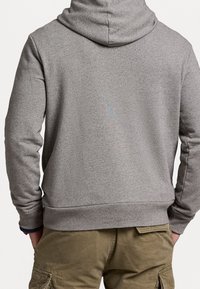 Grå pullover-hoodie med texturerad yta, med en kenguruficka och ribbade muddar, visad bakifrån mot en neutral bakgrund.