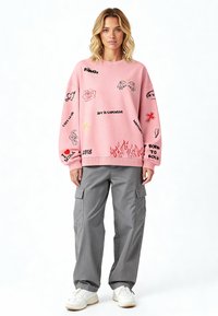 Felpa oversize rosa con vari grafiche ricamate multicolori; abbinata a pantaloni cargo grigi e sneakers bianche.
