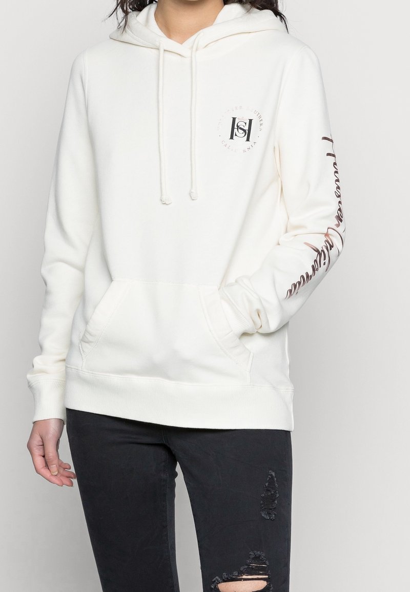 Person, der einen weißen Hoodie mit Fronttasche und schwarze kaputte Jeans trägt, mit einer Hand in der Tasche, teilweise sichtbarer Oberkörper.