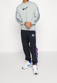 Man som bär en grå Nike-tröja med svart swoosh, svarta träningsbyxor med blå och vinröda ränder, samt vita Nike-sneakers med blå swoosh.