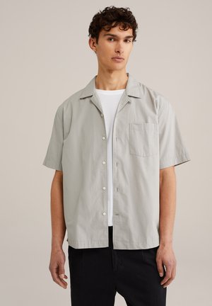 Light gray short-sleeve shirt in a smooth fabric, featuring a button-down front, pointed collar, and a chest pocket. Worn over a white t-shirt.