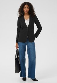 Fekete blézeres nő, fehér csipkebetétes felsőben, kék bővülő nadrágban, fekete hegyes sarkú cipőben, és egy fekete táskával a kezében.