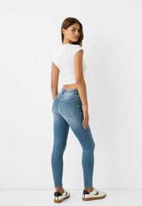 Kék skinny farmernadrág magas derékkal, egy testhez simuló fehér felsővel és sportcipővel párosítva. A farmernadrág klasszikus öt zsebes kialakítással rendelkezik.