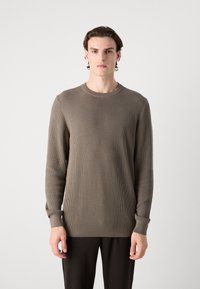 Jersey marrón de manga larga con cuello redondo y puños ajustados, tejido texturizado, corte recto, combinado con pantalones oscuros.