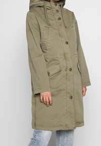 Oliwkowy, średniej długości płaszcz z kapturem, z zapięciem na guziki, dwoma przednimi kieszeniami i długimi rękawami. Materiał wydaje się trwały i teksturowany.
