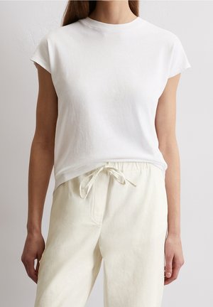 Femme portant un haut blanc uni à manches courtes et un pantalon beige clair à cordon, sur fond neutre.