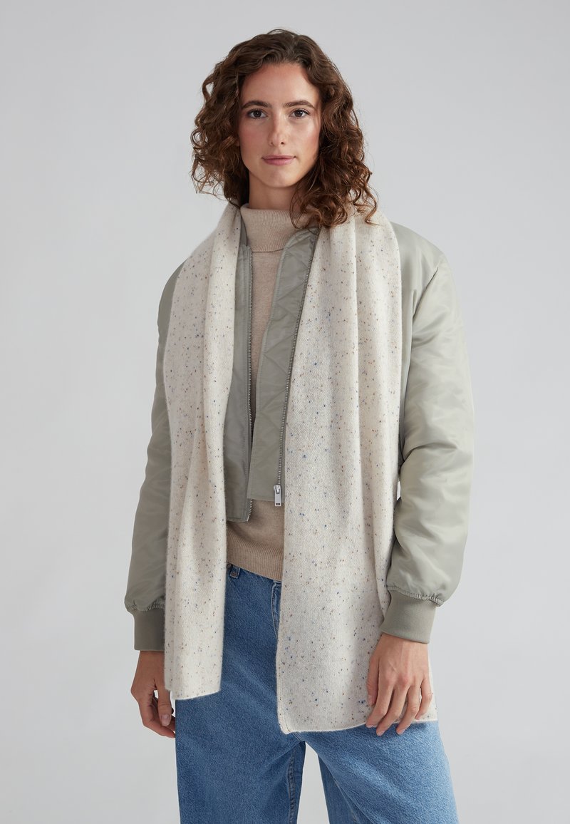 Veste bomber gris clair avec des poignets élastiques, pull beige et écharpe blanche à motifs colorés. Un jean en denim décontracté complète le look.