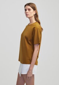 Senffarbenes T-Shirt mit kurzen Ärmeln, runder Ausschnitt, lockere Passform, weicher Stoff, kombiniert mit weißen Shorts, Modell im Profil vor neutralem Hintergrund dargestellt.