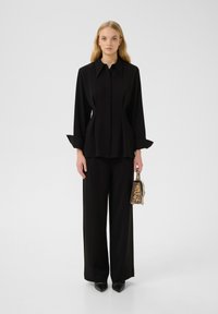 Camicia nera su misura con colletto a punta, vita plissettata e maniche lunghe. Abbinata a pantaloni neri a gamba larga e una piccola borsa a stampa leopardata.