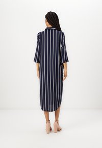 Abito blu navy con strisce verticali bianche, maniche a tre quarti e colletto. Design fluido, lunghezza a metà polpaccio con uno spacco laterale.