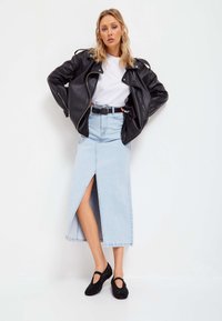 Giubbotto di pelle nera da motociclista, t-shirt bianca, gonna lunga in denim azzurro chiaro con spacco frontale e scarpe nere decorate.