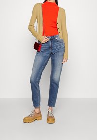 Cardigan beige à côtes porté sur un haut col montant rouge, associé à un jean bleu et des chaussures marron à lacets avec semelles épaisses. Petit sac à main rouge inclus.