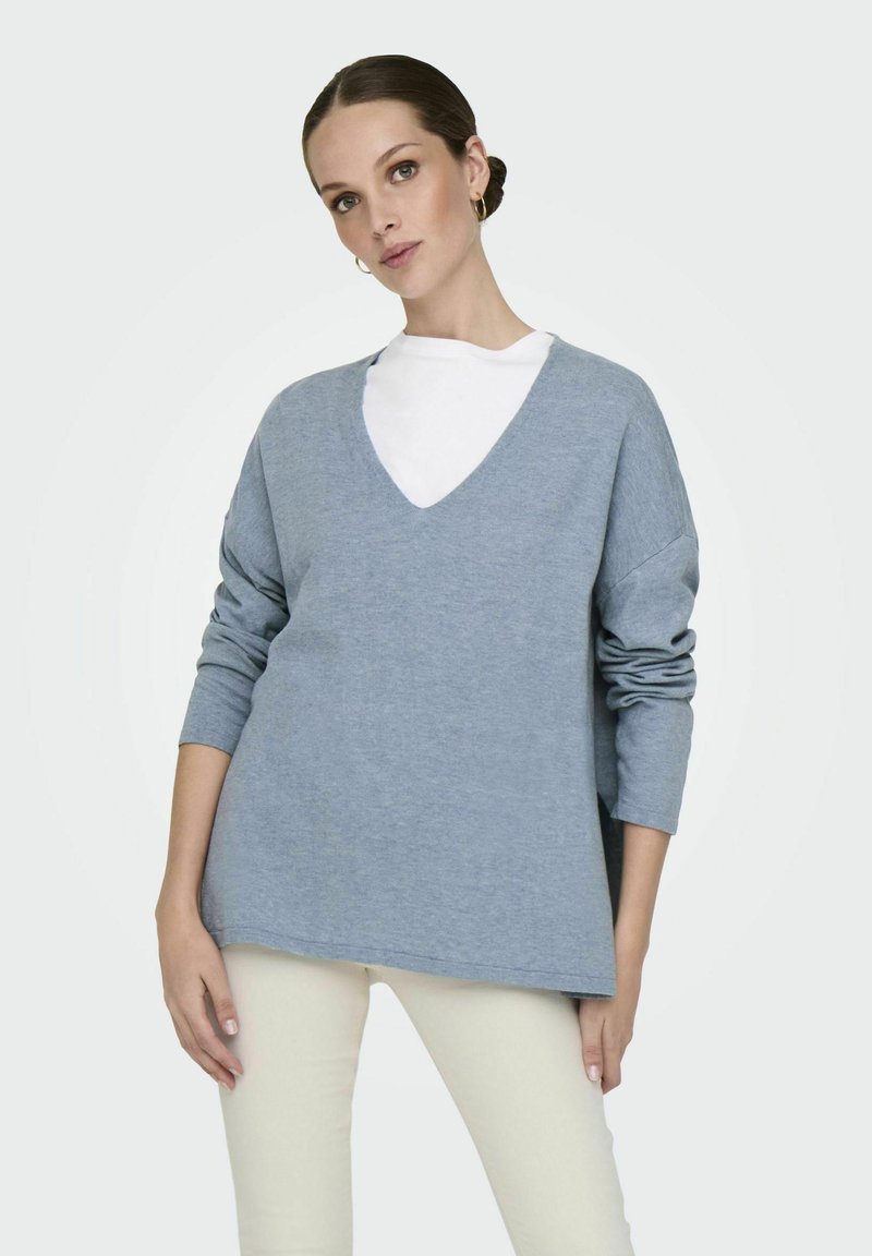 Maglione azzurro chiaro, dalla vestibilità ampia, con scollo a V, maniche lunghe e polsini arrotolati, indossato sopra una maglietta bianca, abbinato a pantaloni crema.