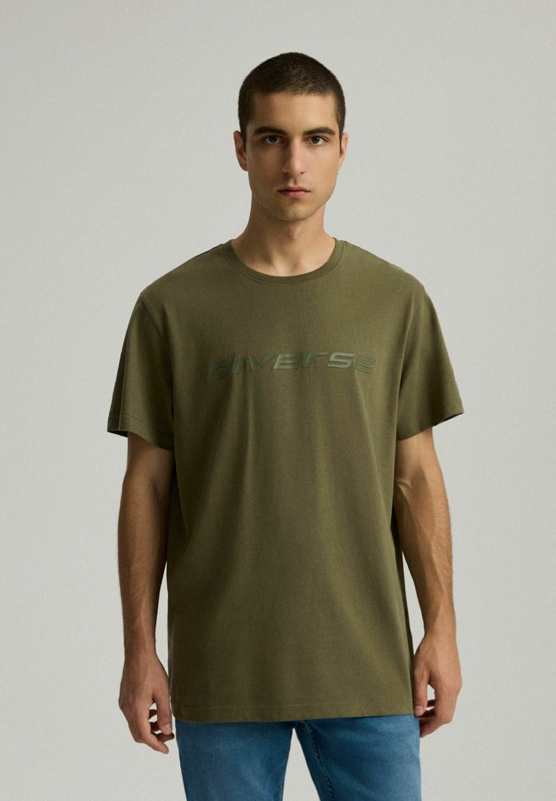 Oliwkowa bawełniana koszulka z krótkimi rękawami, z okrągłym dekoltem, z napisem "Diverse" w ciemniejszym odcieniu zieleni na wysokości klatki piersiowej.