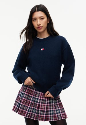 Jeune femme aux longs cheveux foncés portant un pull bleu marine et une jupe plissée à carreaux rose sur fond blanc.