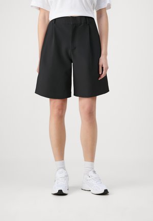 Personne portant un short noir mi-longueur, une chemise blanche à manches courtes, des chaussettes blanches et des chaussures de sport blanches, debout sur fond blanc.