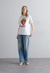 T-shirt bianca a maniche corte con una grande grafica colorata di fragola e la scritta "BALLY" in testo blu; abbinata a jeans ampi di un leggero blu.