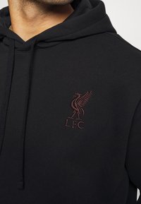 Hoodie noir en mélange de coton avec une poche kangourou. Présente un logo L.F.C. brodé en rouge foncé sur la poitrine. Capuche réglable avec cordon.