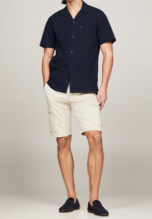 Mann trägt marineblaues kurzärmeliges Hemd mit Knöpfen, beige Cargo-Shorts mit Taschen und marineblaue Slipper, steht mit Händen in den Taschen.