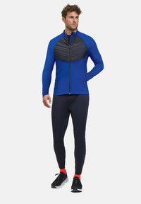 Blauwe sportjack met een zwart gewatteerd paneel, rits aan de voorkant en zijzakken, gecombineerd met zwarte aansluitende broek en zwarte schoenen met rode accenten.