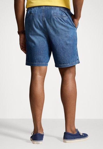 Shorts en denim de couleur bleu moyen avec une coupe décontractée, une taille élastique, des poches latérales et deux poches arrière. Portés avec des chaussures à enfiler bleu marine.