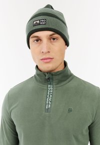 Zeleni polarni pulover s visokim ovratnikom, s četvrtastim zipom i brendiranim trakama. Pleteni zeleni beanie s prugastim uzorkom i logotipom.