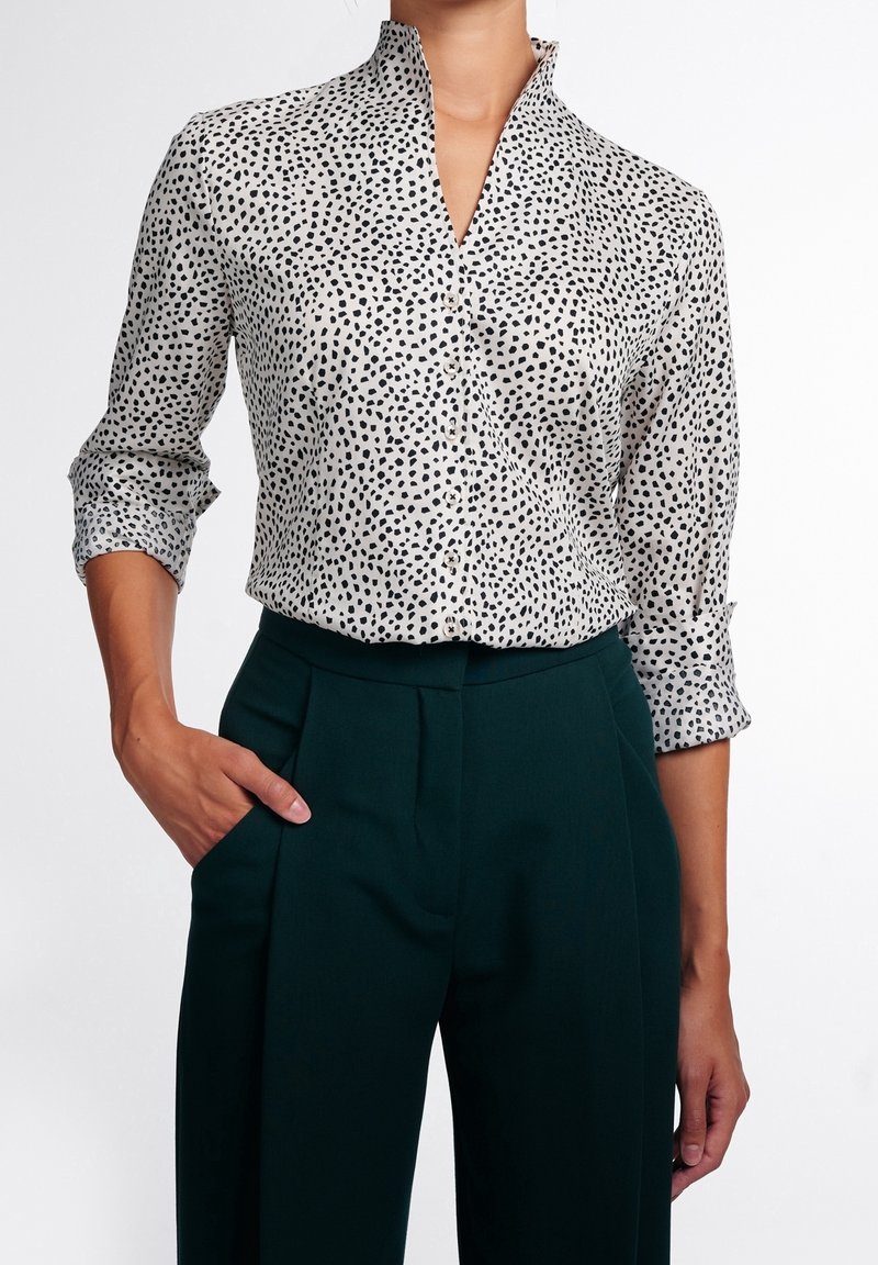 Femme portant un chemisier blanc à manches longues avec des taches noires et un pantalon taille haute vert foncé, une main dans la poche.