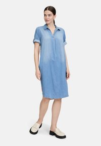 Niebieska sukienka koszulowa z denimu z kołnierzykiem, krótkimi podwiniętymi rękawami i prostym krojem. Posiada zaokrąglony dół i widoczne detale po bokach. Czarne buty na platformie.