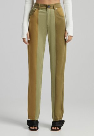 Dwukolorowe, proste spodnie z wysokim stanem w odcieniach oliwkowym i musztardowym, noszone z czarnymi sandałami z odkrytymi palcami oraz białą, długorękawową, krótka bluzką.