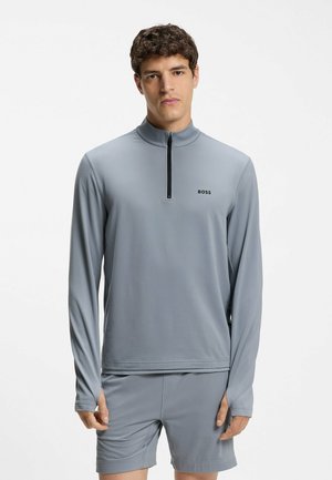 Giovane uomo che indossa un maglione grigio chiaro a maniche lunghe con una corta zip e pantaloncini coordinati, in piedi davanti a uno sfondo chiaro e semplice.