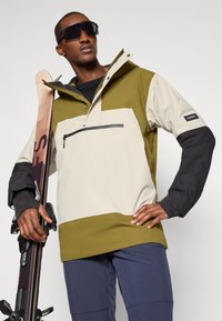 Hiihtotakki vaaleassa beigeissä, oliivinvihreissä ja mustissa väreissä. Varustettu puolivetoketjulla, säädettävällä hupulla ja vetoketjullisella rintataskulla. Sileä kangaspinta.