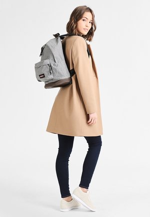 Mochila gris con un asa superior y un bolsillo frontal, llevada sobre un abrigo beige. El modelo lleva vaqueros oscuros y zapatillas claras. Detalle sutil de textura.
