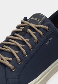 Ναυτικό μπλε αθλητικό παπούτσι Geox με μπεζ πλεκτά κορδόνια περασμένα από μεταλλικά κορδόνια, με λευκή ελαστική σόλα και επωνυμίες.