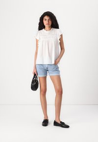 Blouse blanche à manches courtes avec des bords volants, en tissu texturé, col rond, assortie à un short en denim clair, ainsi qu'un sac à main noir et des chaussures noires.