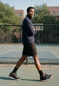 Jeune homme en veste sombre et short marchant le long d'une clôture en grillage près d'un terrain de basket en plein air par une journée ensoleillée.