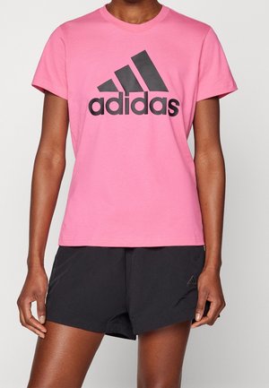 Osoba nosząca różowy t-shirt Adidas z czarnym logo oraz czarne sportowe spodenki Adidas, stojąca na tle jednolitego jasnego tła.