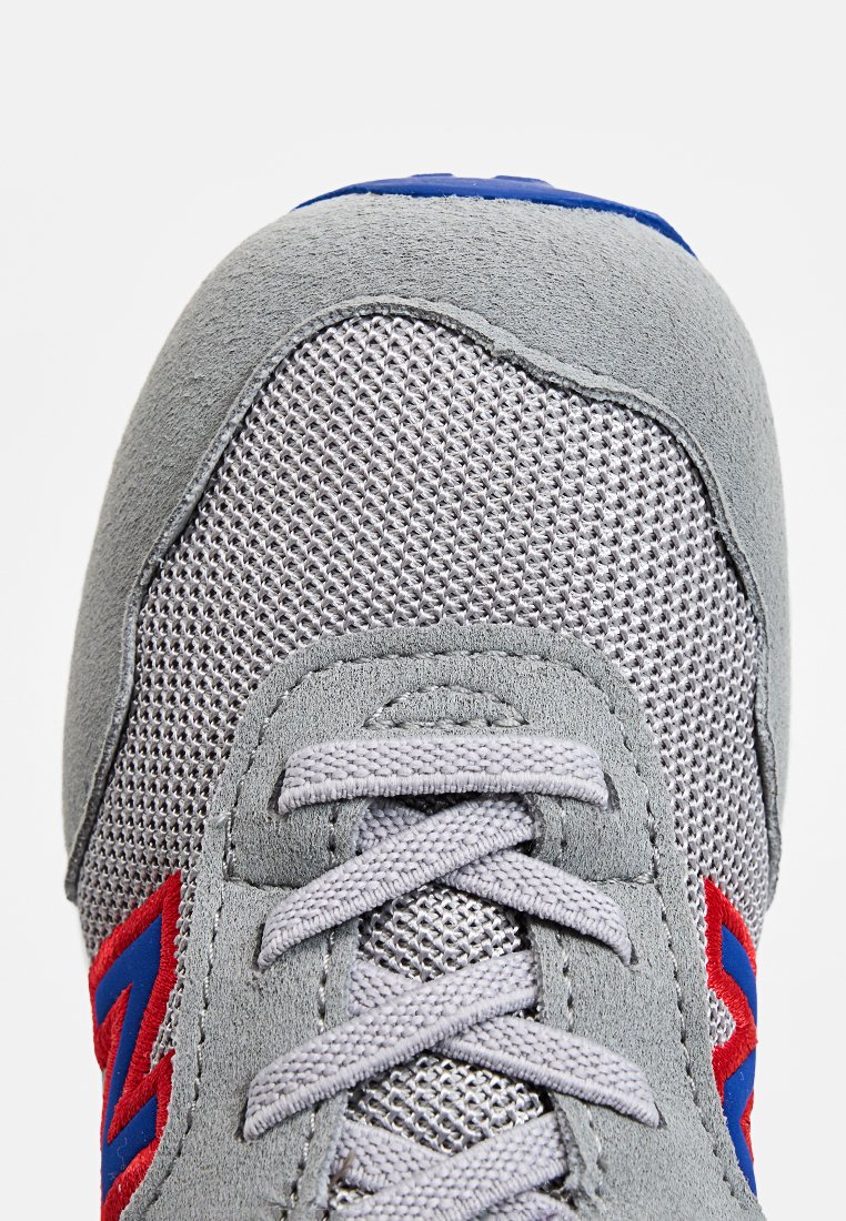 Zapatilla gris con materiales de malla y ante, con acentos rojos y detalles azules en la parte superior. Incluye un diseño de cordones planos.