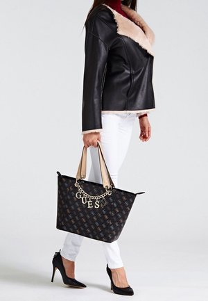 Femme tenant un sac fourre-tout noir de créateur avec chaîne dorée et lettres, portant une veste à col en fourrure noire, un pantalon blanc et des talons noirs.