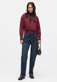 Bordo kapucinski s pulover, s oblečeno bandano okrog vratu, v kombinaciji z temno modrimi ravnimi hlačami, črno studded torbico in črnimi čevlji.