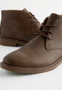 Botas de tobillo de cuero marrón con una textura lisa, punta redonda y diseño con cordones. Costuras en contraste y un tacón bajo apilado.