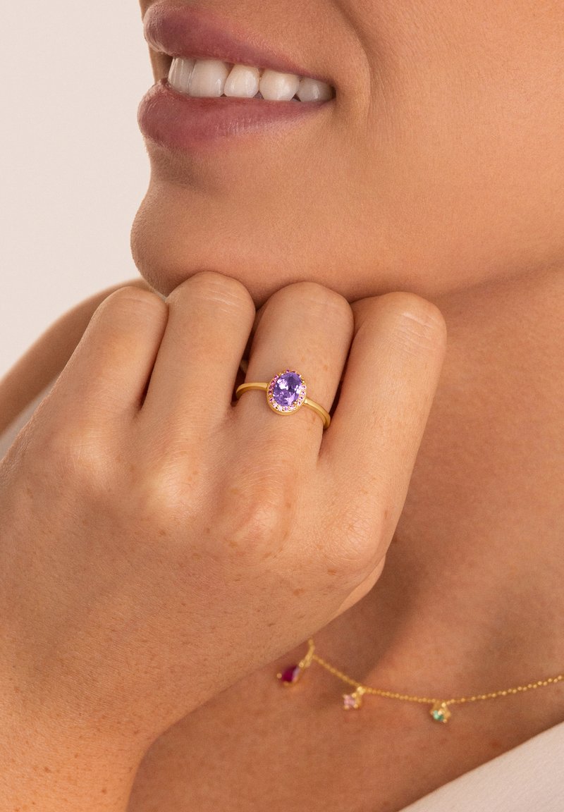 Main avec une bague en or ornée d'une pierre précieuse ovale violette près de lèvres souriantes, et un délicat collier en or avec des breloques en pierres précieuses colorées.