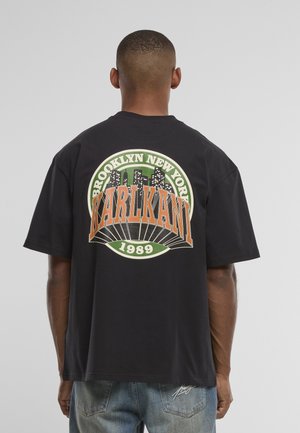 Mężczyzna w czarnej, dużej koszulce z logo "Brooklyn New York Karl Kani 1989" i wzorem panoramy miasta na plecach.