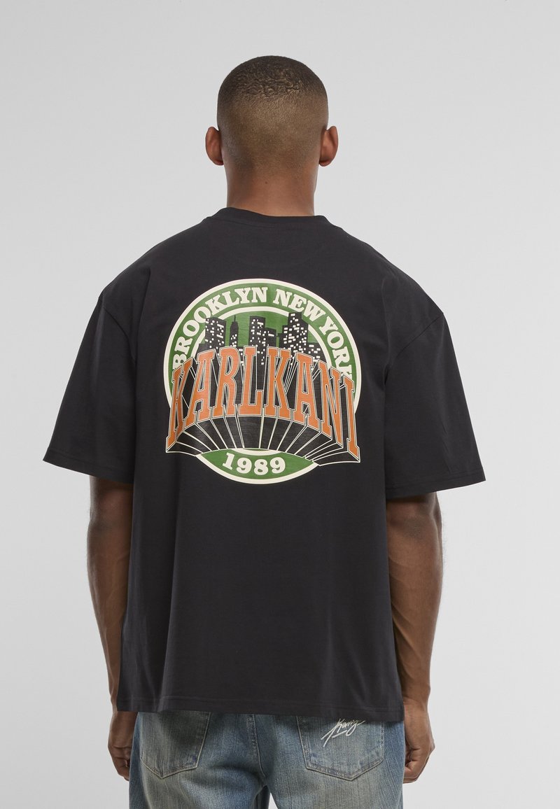 Άνδρας φοράει μαύρο oversized T-shirt με το λογότυπο "Brooklyn New York Karl Kani 1989" και σχεδιασμό του ορίζοντα της πόλης στην πλάτη.