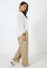 Un haut oversized en tricot ouvert blanc avec des manches chauve-souris, associé à un pantalon large beige et des baskets blanches, sur fond uni.