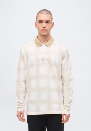 Άνδρας που φοράει μπλουζάκι πόλο Nike Golf με μακριά μανίκια σε μπεζ και λευκό χρώμα, με μπεζ γιακά, στέκεται μπροστά σε απλό ανοιχτό γκρι φόντο.