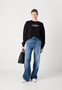 Felpa nera con la parola "BOSS" in bianco, abbinata a jeans a gamba larga blu chiaro. Una borsa a tracolla nera è tenuta in una mano.