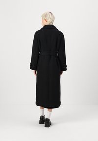 Casaco longo preto feito de um tecido suave, com cintura cingida por um cinto, grande gola e punhos com botões. Design limpo e minimalista.