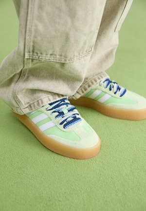 Personne portant un pantalon beige et des baskets vert clair avec des lacets bleus et des semelles en gomme, debout sur un tapis vert clair.